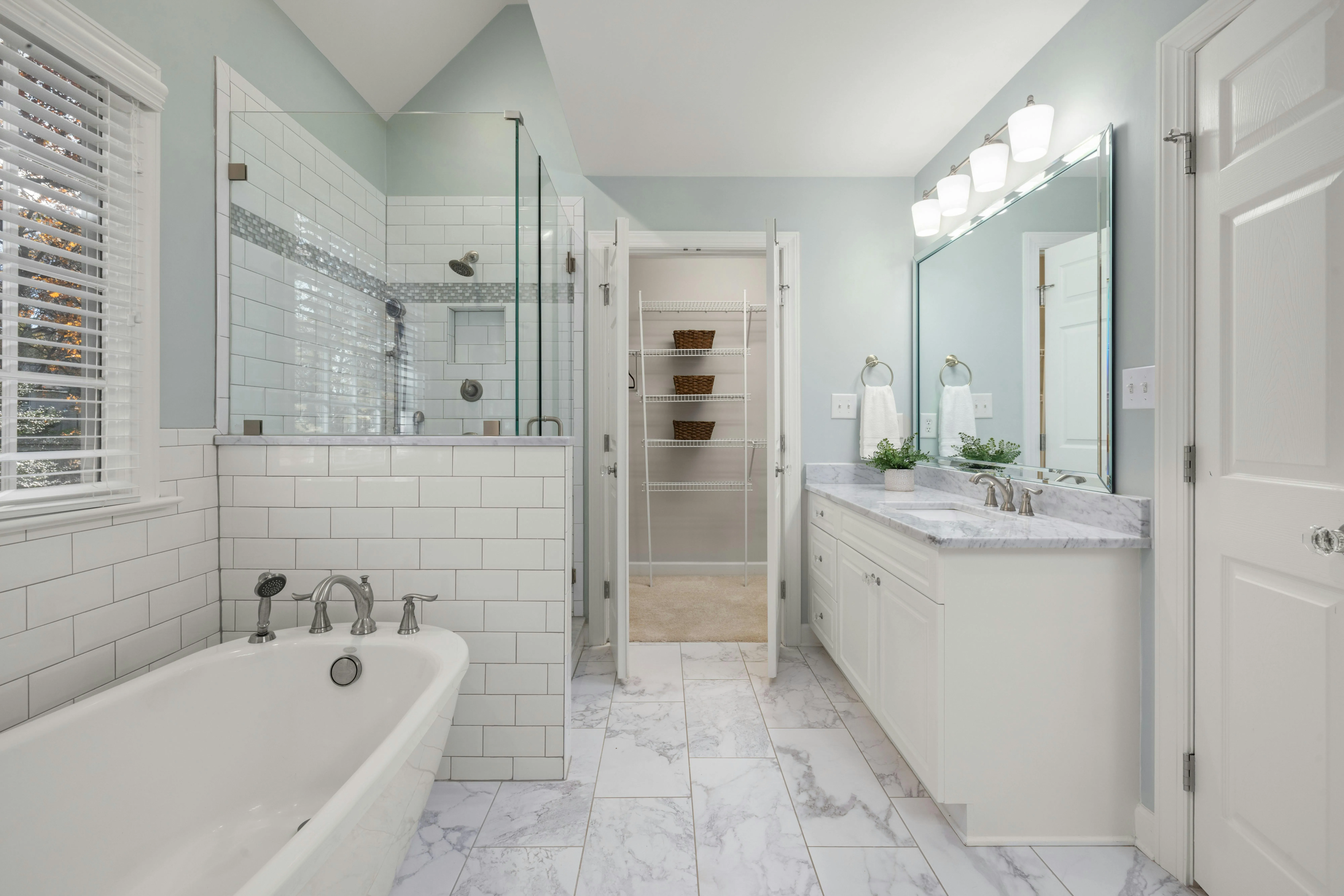 Bright bathroom with a white bathtub, glass-enclosed shower, marble floor tiles, and a vanity with a large mirror and double sinks.