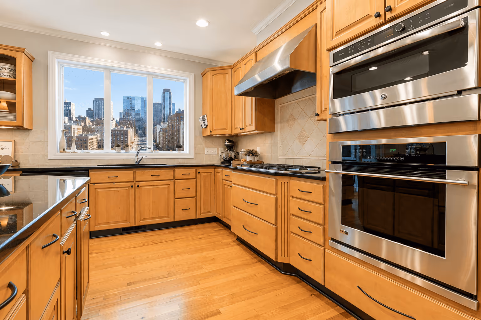 Modern kitchen with wooden cabinets, stainless steel double oven, black granite countertops, hardwood floor, and a large window showing a city skyline.