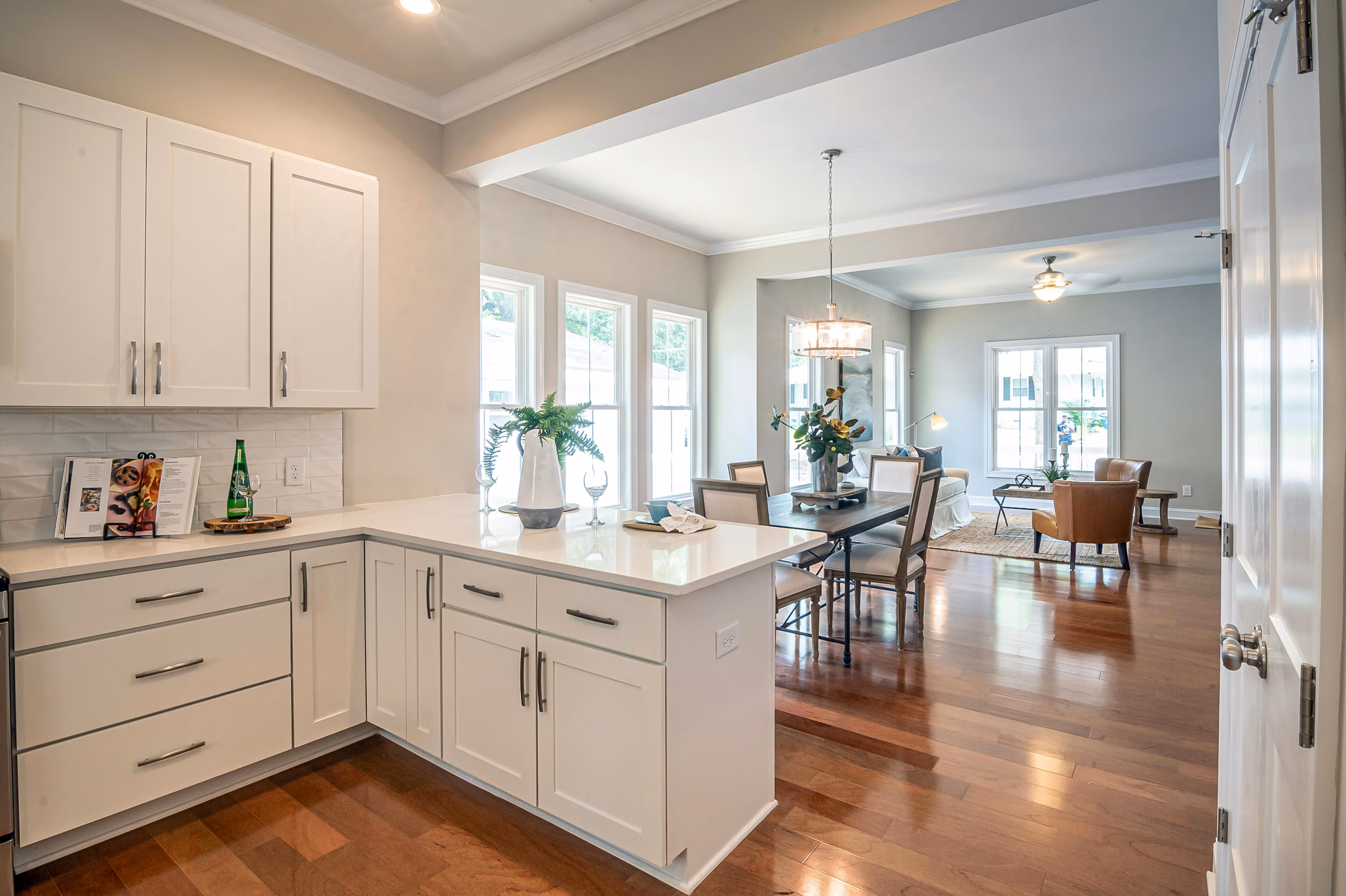 Open kitchen with white cabinets and countertops, overlooking a dining area with a wooden table and chairs, and a living room with seating and large windows.