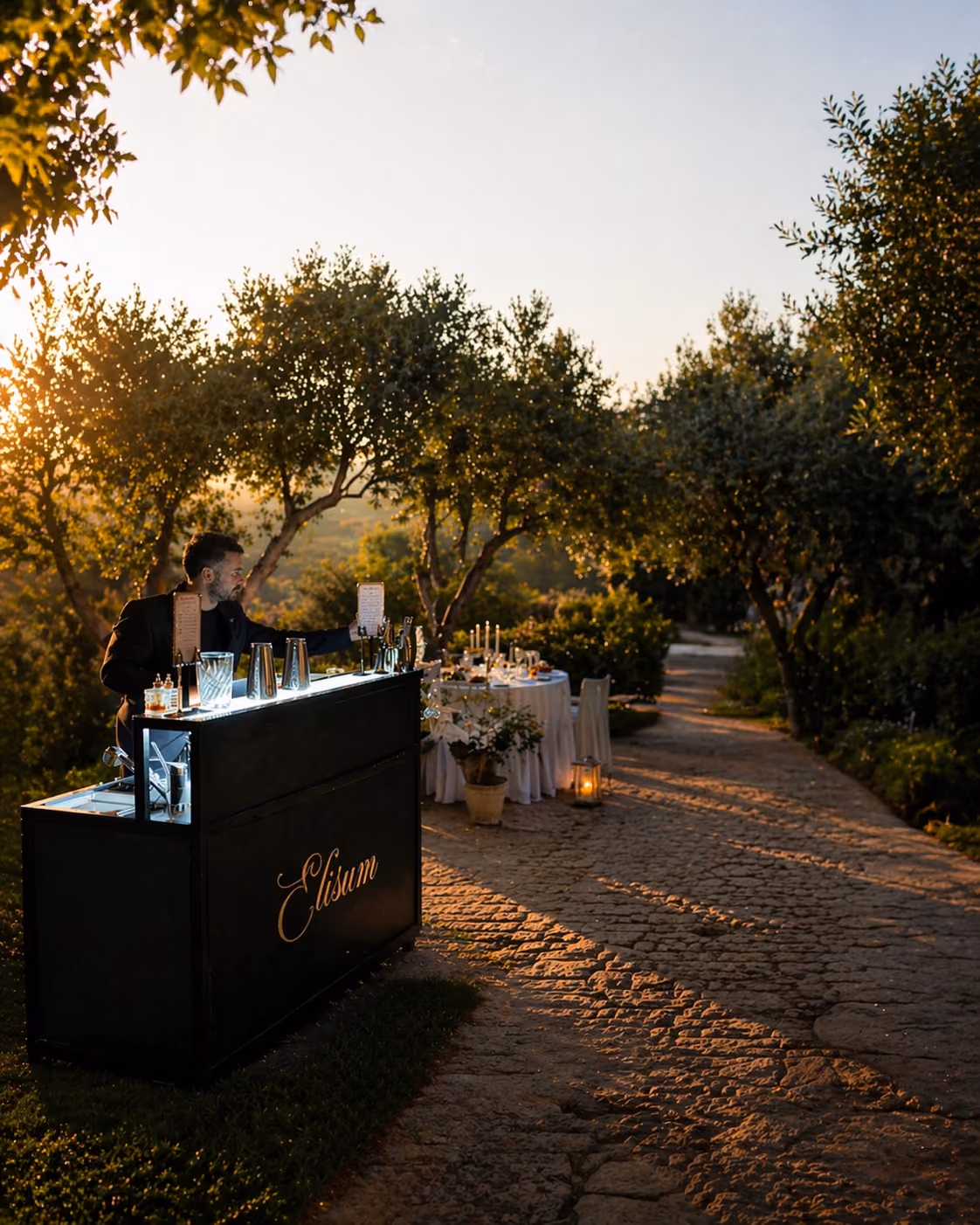 Man tending an outdoor bar at sunset with a decorated round table and trees nearby.