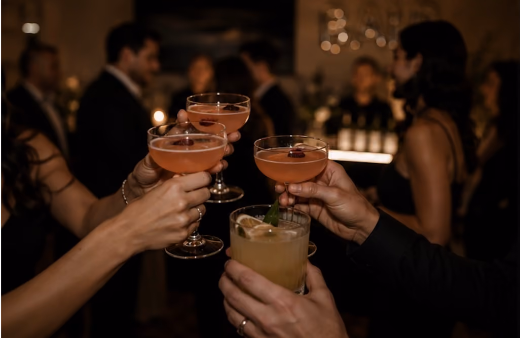 Hands holding cocktails clinking glasses at a dim party with people mingling in the background.