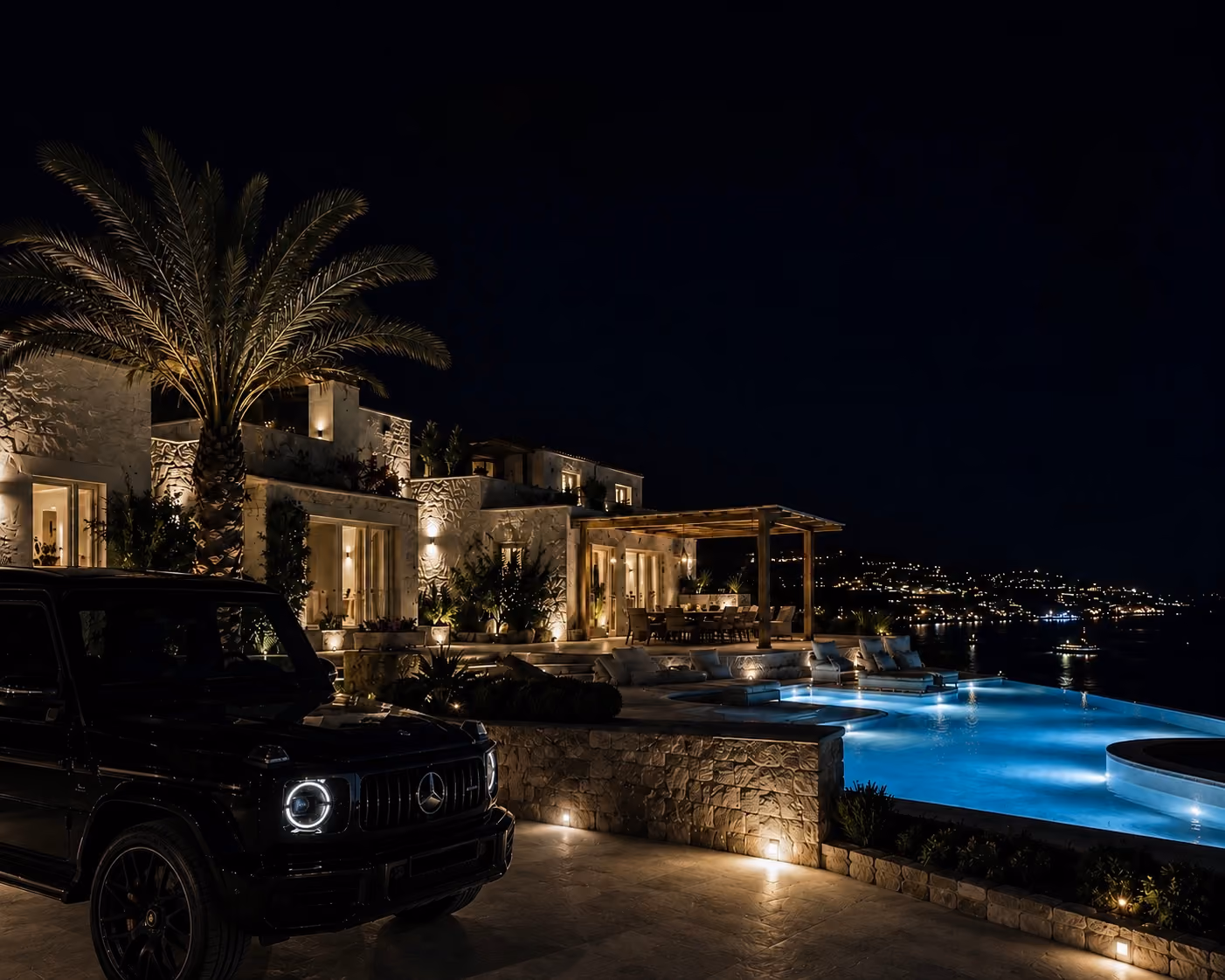 Luxury villa at night with lit infinity pool, outdoor dining area, palm tree, and a black Mercedes G-Class parked outside.