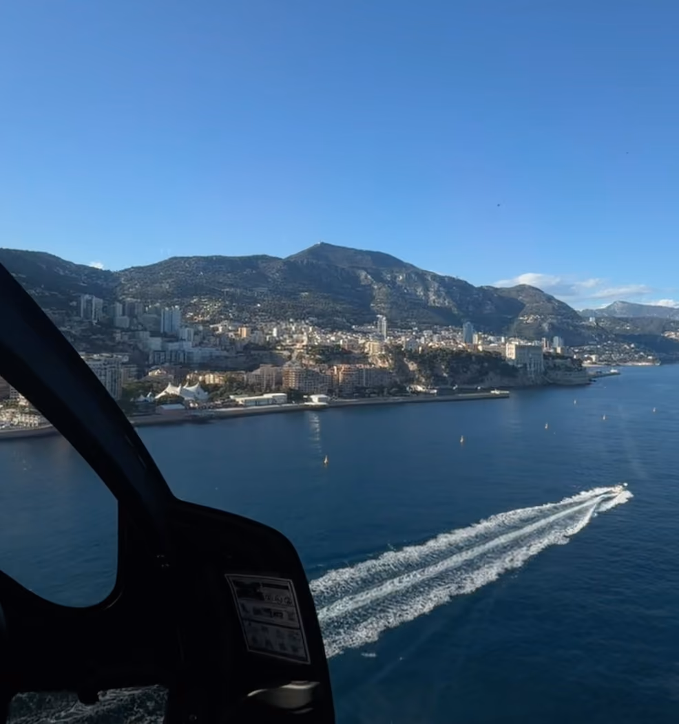 Aerial view of a coastal city with mountains, blue sky, and a boat leaving a white trail in the water.