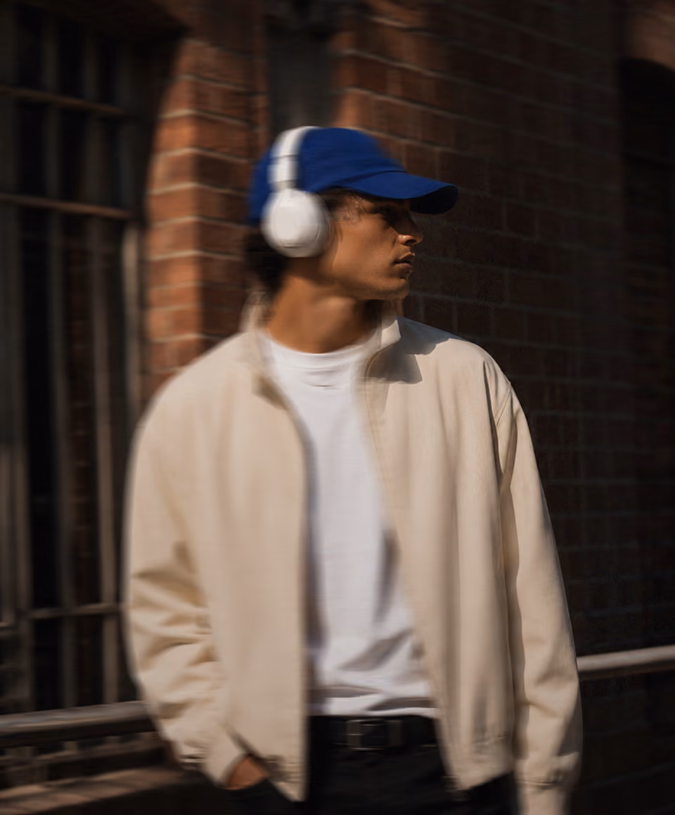 Person wearing white headphones, blue cap, white jacket, and white shirt standing against a brick wall.