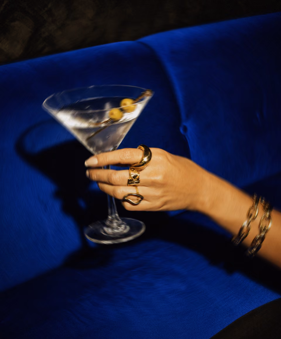 Hand with gold rings and bracelets holding a martini glass with two olives on a blue couch.