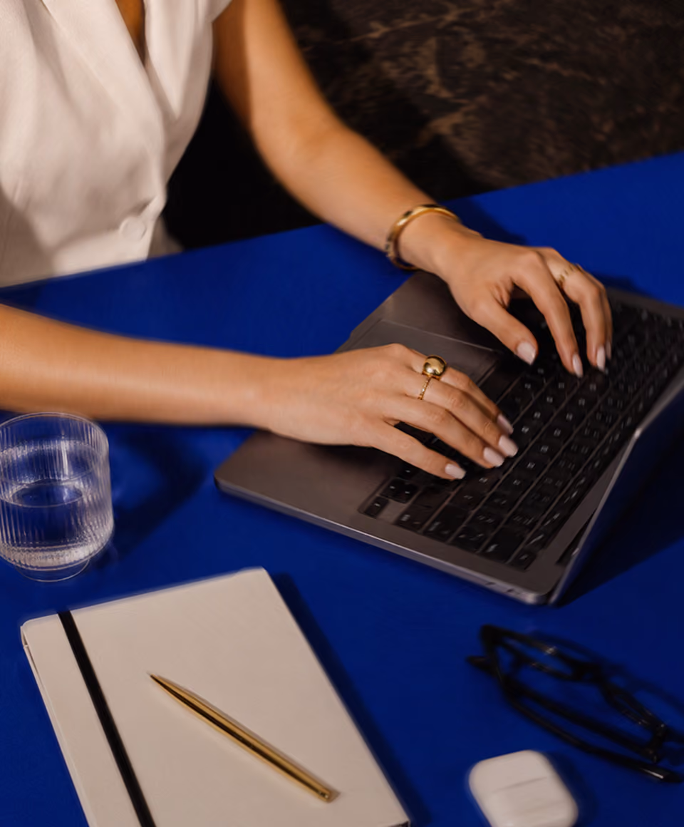 Person typing on laptop at blue desk with notebook, pen, glasses, and glass of water nearby.