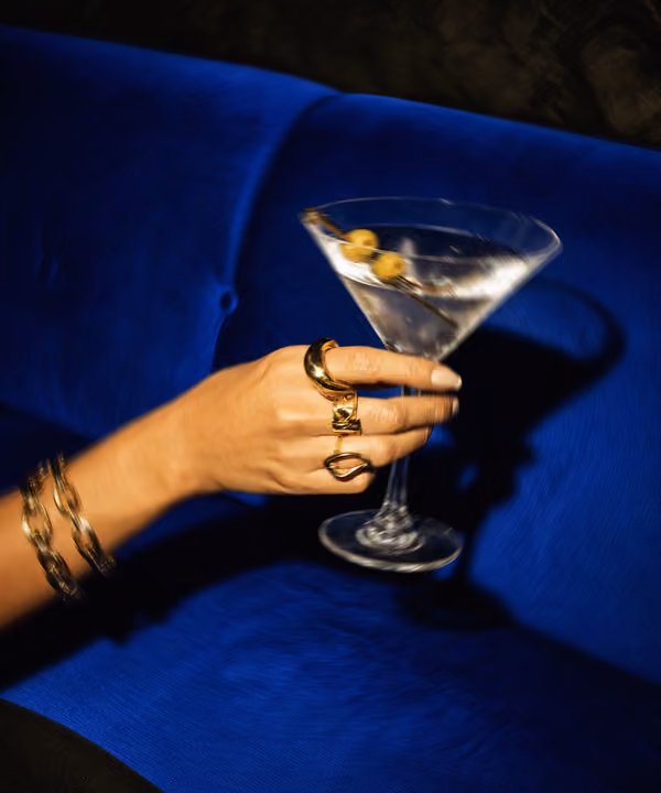 Hand with gold rings and bracelets holding a martini glass with olives against blue background.