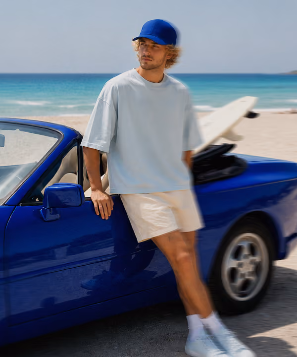 Man in a blue cap and light shirt leaning on a blue convertible car parked on a beach with surfboard.