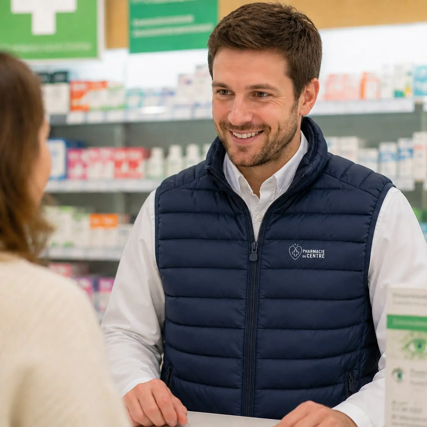 Pharmacien souriant en gilet bleu discutant avec une cliente à la pharmacie.