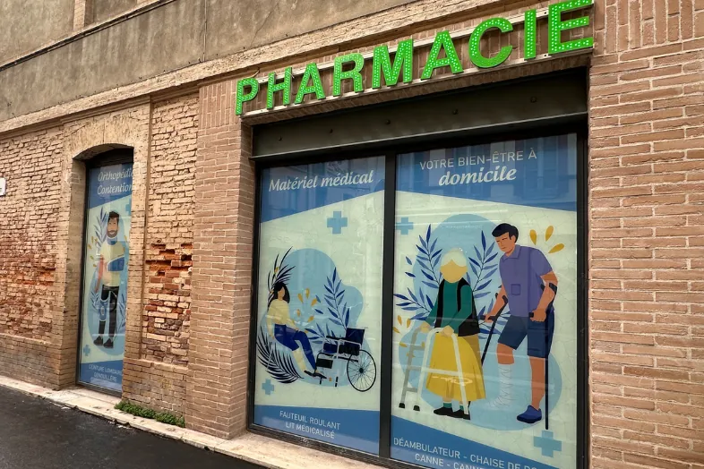 Façade d'une pharmacie avec enseigne verte et affiches sur matériel médical et soins à domicile.
