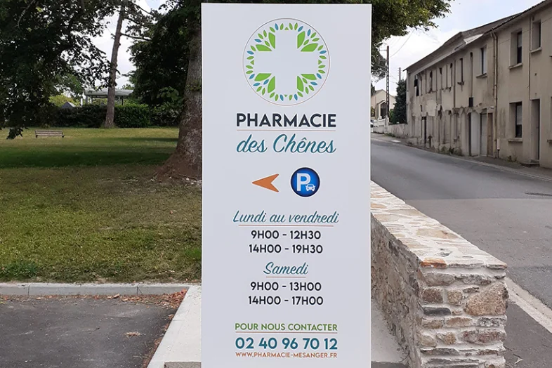 Panneau de la Pharmacie des Chênes avec horaires d'ouverture et contact en rue près d'un parc.