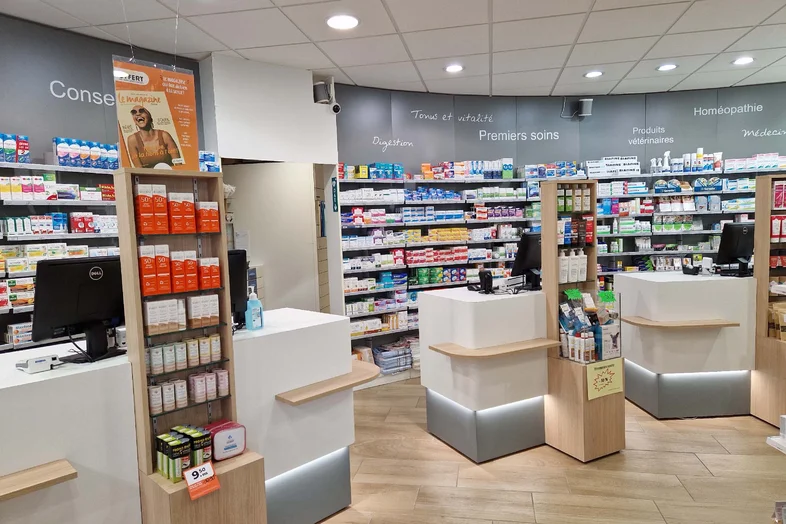Intérieur d'une pharmacie avec plusieurs comptoirs blancs et étagères remplies de médicaments et produits.