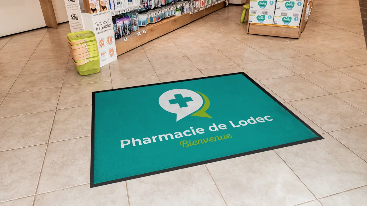 Tapis vert avec logo et texte 'Pharmacie de Lodec Bienvenue' devant des rayons en pharmacie.