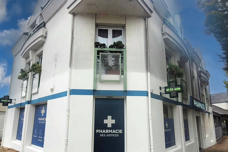 Façade d'une pharmacie blanche avec une croix verte et des plantes aux fenêtres.