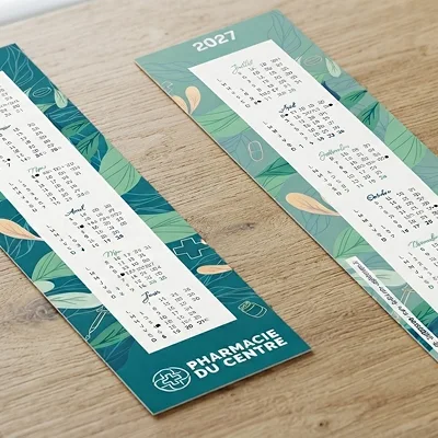 Deux calendriers 2027 verts avec motifs végétaux posés sur une surface en bois, marqués Pharmacie du Centre.