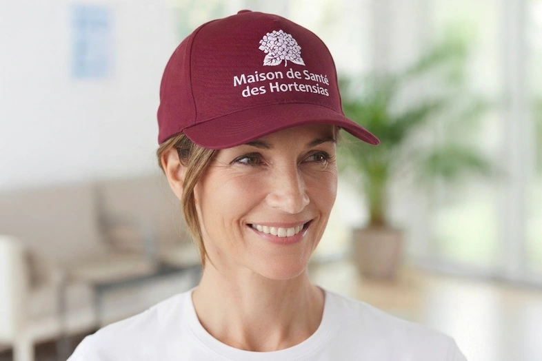 Femme souriante portant une casquette bordeaux avec logo et texte Maison de Santé des Hortensias.