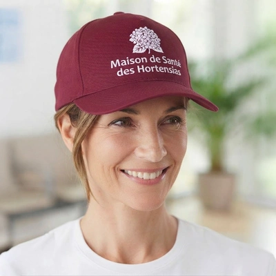 Femme souriante portant une casquette bordeaux avec un logo et texte Maison de Santé des Hortensias.