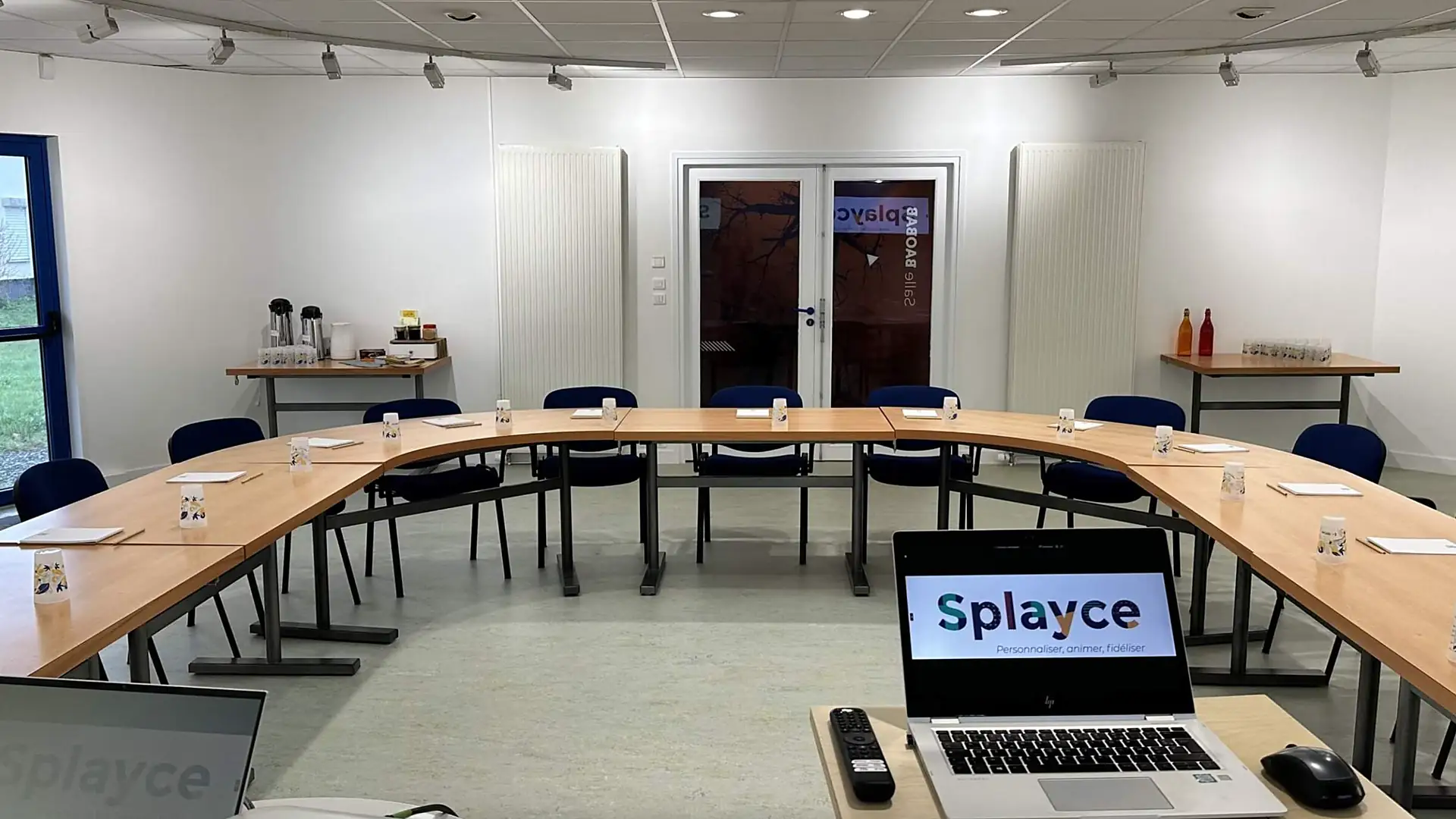 Salle de réunion avec tables en U, chaises bleues, et un ordinateur portable affichant le logo Splayce.