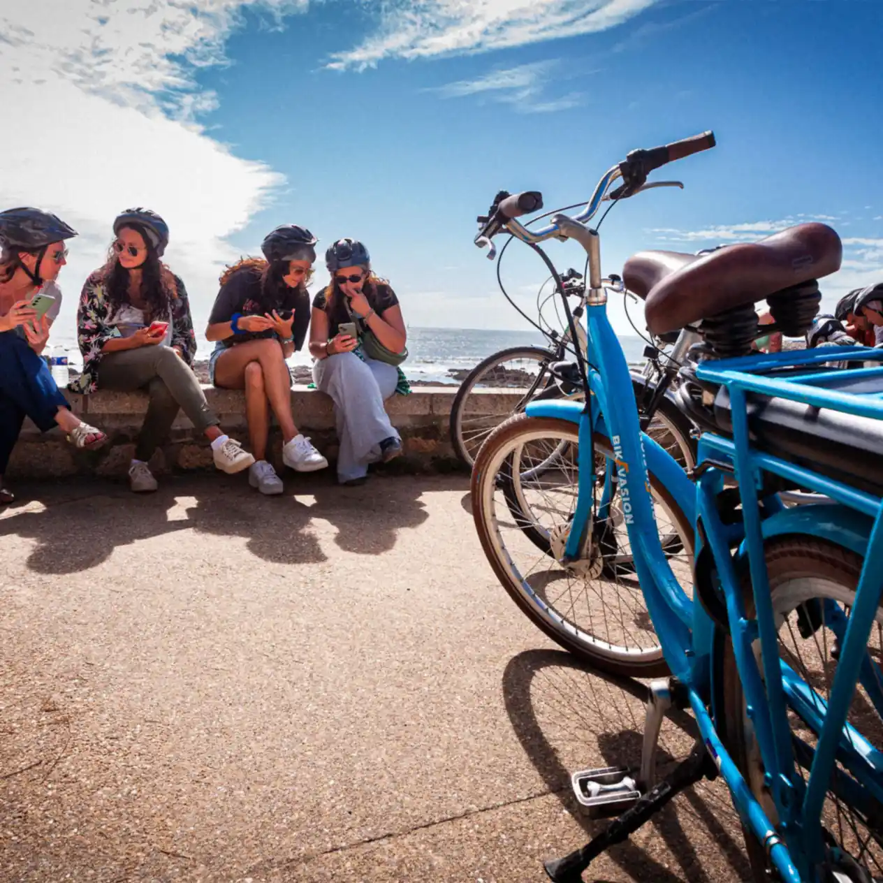 Quatre femmes portant des casques regardant leurs téléphones près de vélos bleus au bord de la mer.