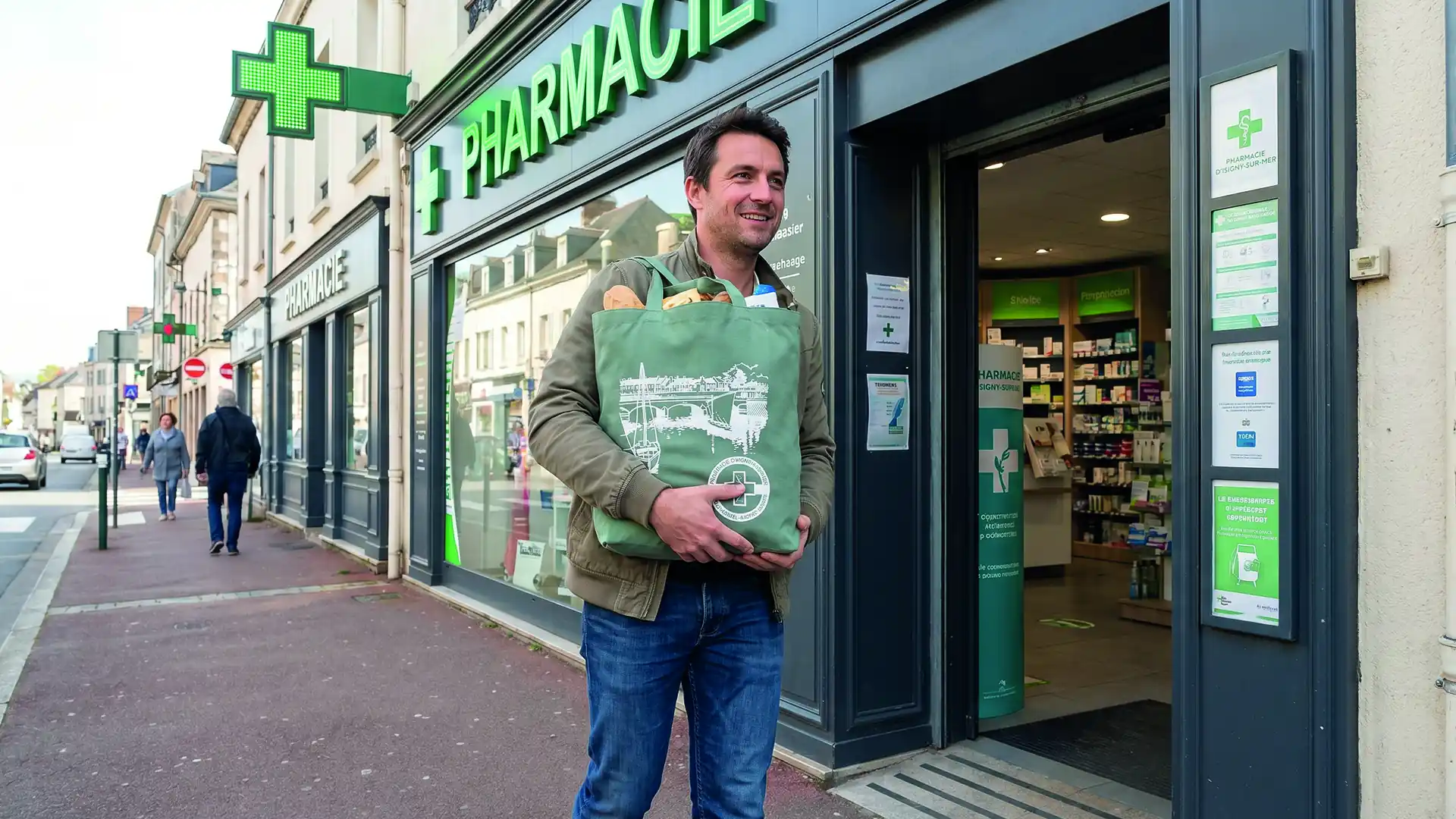Homme souriant sort d'une pharmacie avec un sac réutilisable vert rempli d'achats.