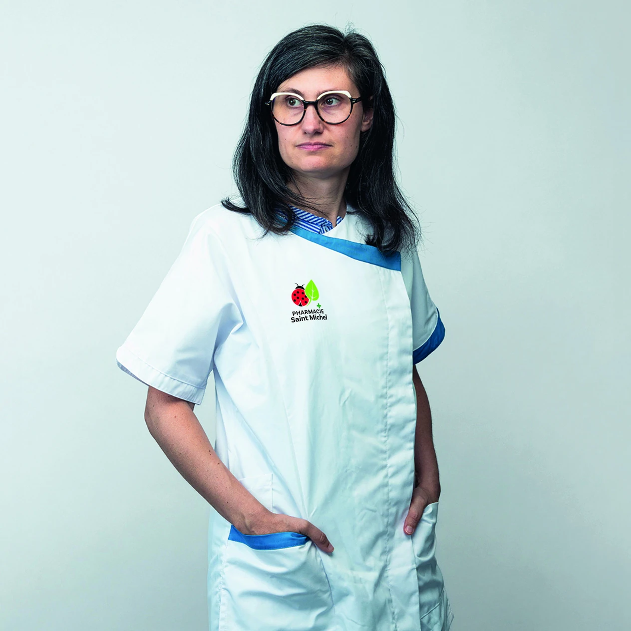 Femme portant une blouse blanche avec logo Pharmacie Saint Michel et bordure bleue, mains dans les poches.