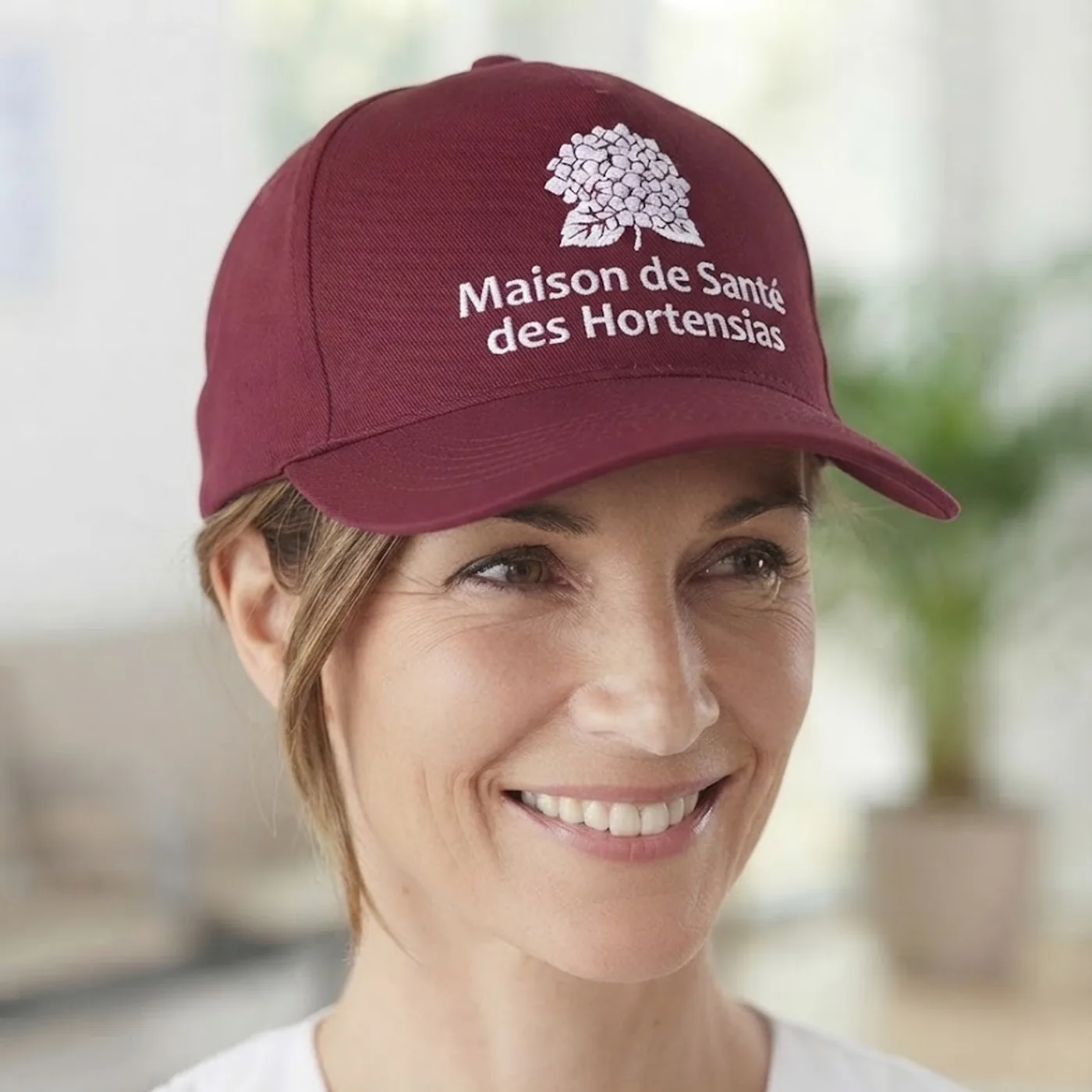 Femme souriante portant une casquette bordeaux avec logo et texte Maison de Santé des Hortensias.