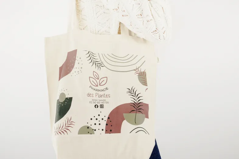 Sac en toile beige avec motifs botaniques et texte Pharmacie des Plantes, numéro et logos réseaux sociaux.