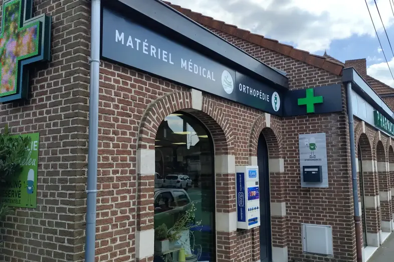 Façade en brique d'une pharmacie avec enseigne 'Matériel médical Orthopédie' et croix verte lumineuse.