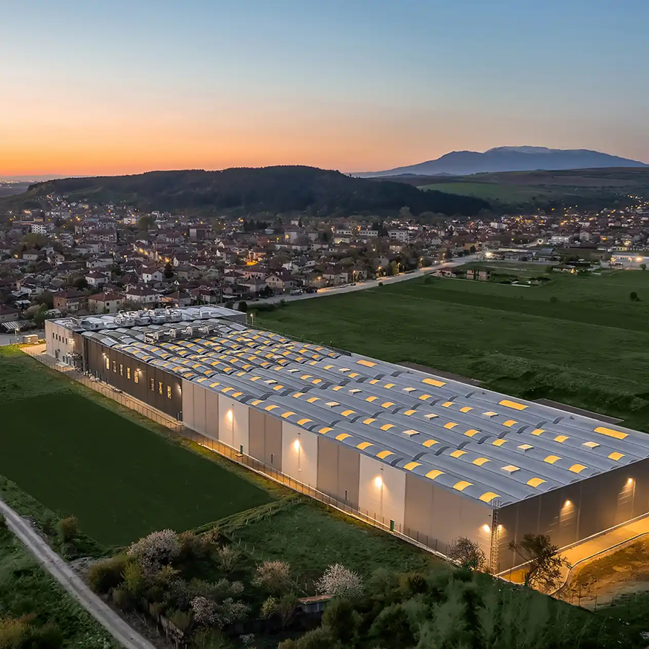 Grand bâtiment industriel éclairé au crépuscule près d'un village et de collines verdoyantes