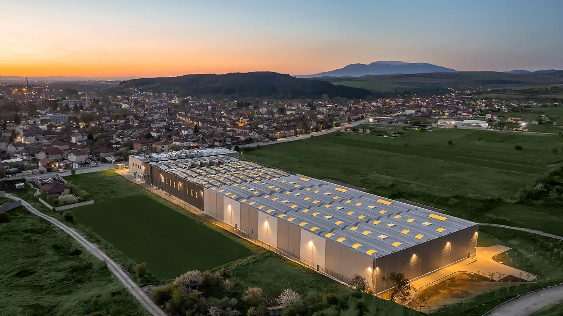 Vue aérienne d'une grande usine éclairée au crépuscule à côté d'une petite ville et des collines.