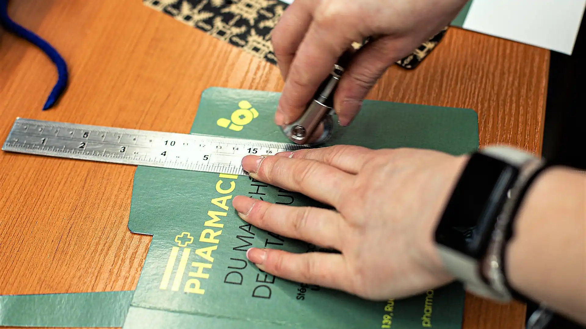 Une personne mesure un carton vert avec une règle métallique sur une table en bois.
