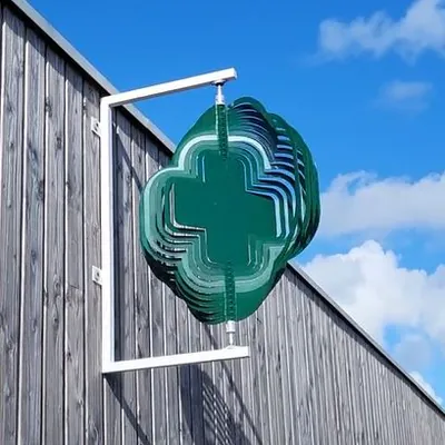 Enseigne croix verte cinétique qui tourne fixée à un mur en bois sous un ciel bleu.