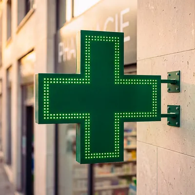 Enseigne lumineuse verte en forme de croix suspendue sur un mur de pharmacie urbaine.
