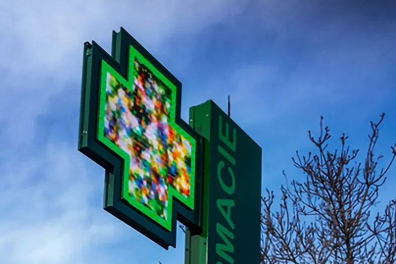 Enseigne lumineuse verte en forme de croix pour une pharmacie avec ciel bleu et branches d'arbre en arrière-plan.