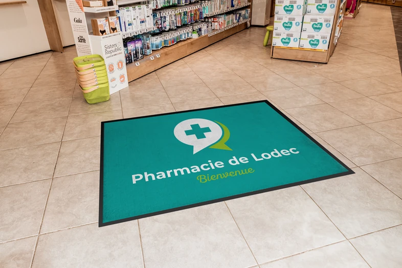 Tapis vert avec logo de pharmacie et texte "Pharmacie de Lodec Bienvenue" sur un sol carrelé.