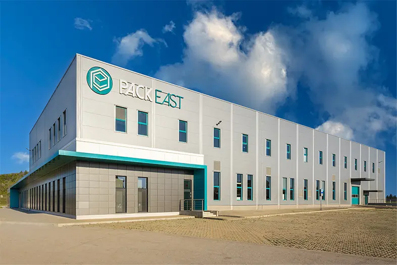 Bâtiment industriel moderne gris avec logo PACK EAST sous ciel bleu partiellement nuageux.