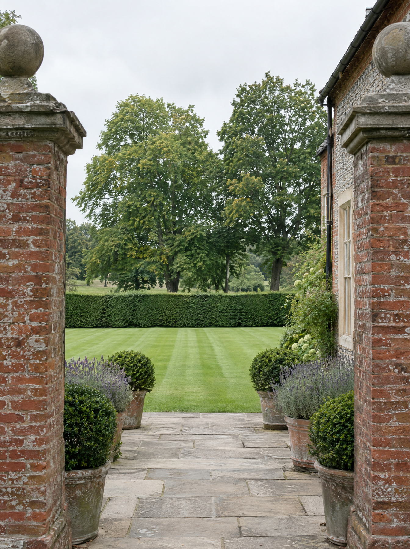 View through brick gate piers along a stone path lined with planted pots towards a striped lawn