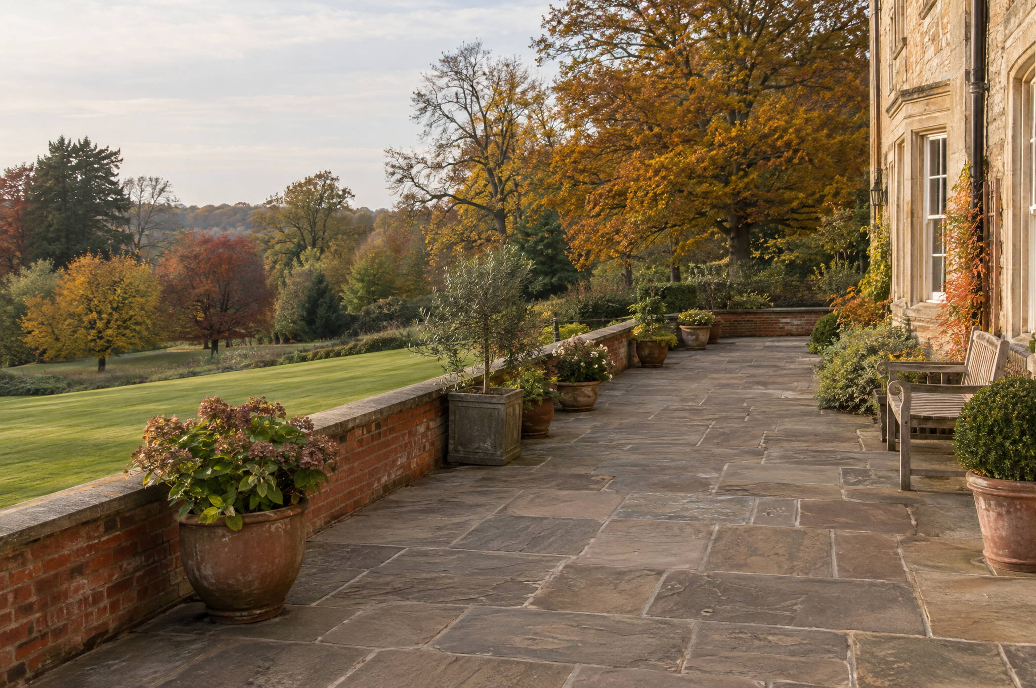 York stone terrace beside a country house in autumn, swept clean of leaves with planted pots and trees turning colour beyond the lawn