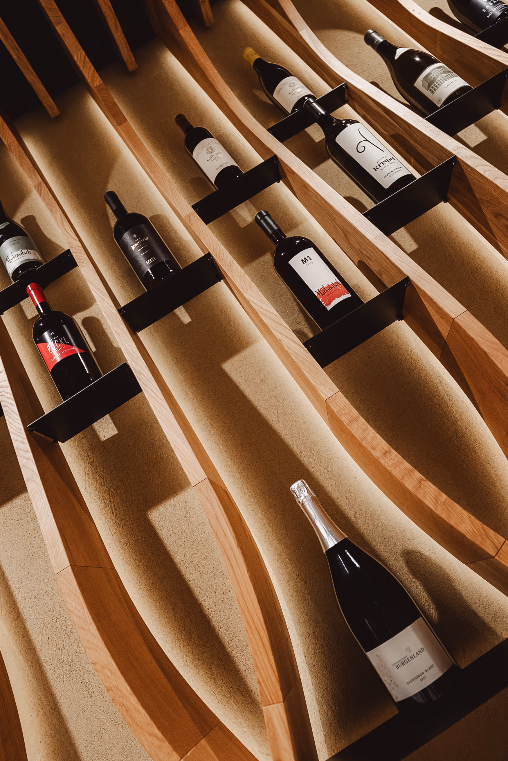 Wall-mounted wooden wine rack with several bottles of wine in angled holders casting shadows on a beige wall.