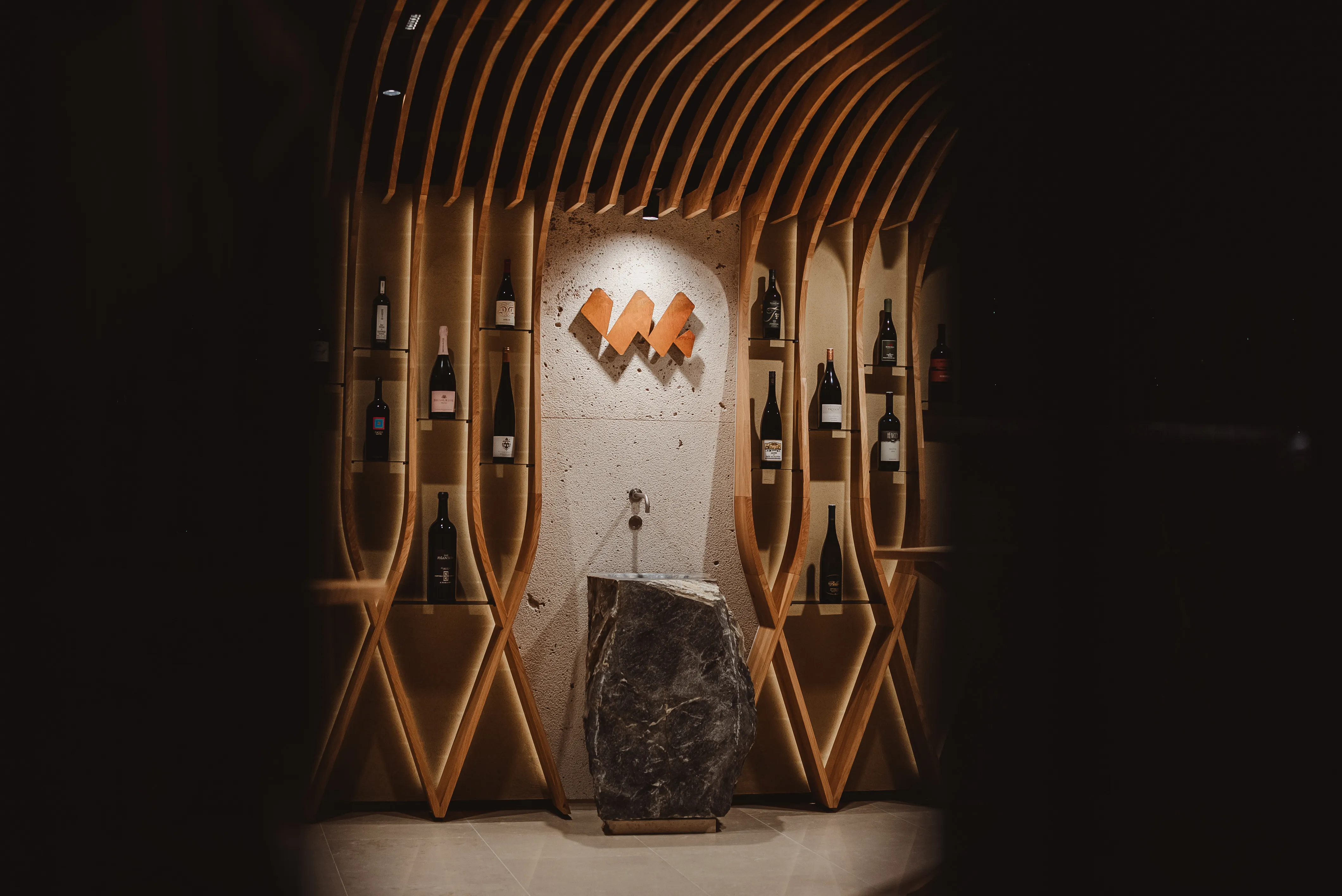 Modern wine display with wooden lattice shelves holding bottles around a stone pedestal and wall logo.