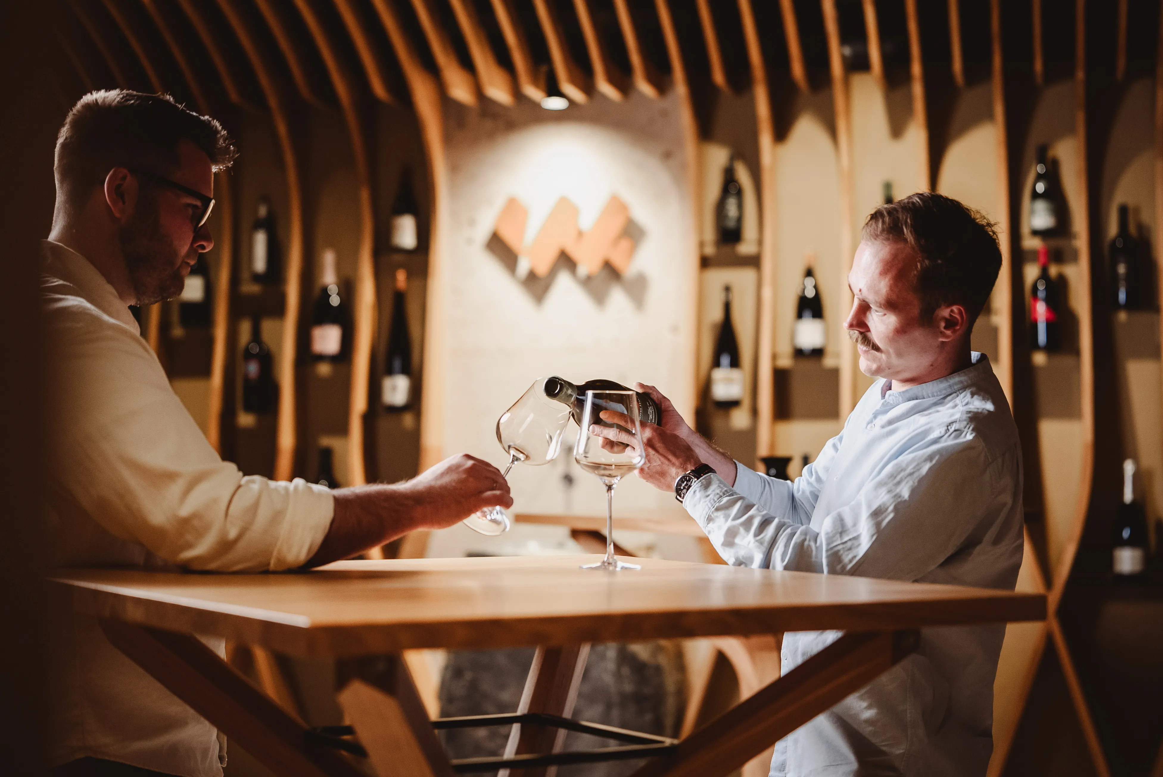 Two men at a wooden table, one pouring white wine into a glass for the other in a modern wine cellar.