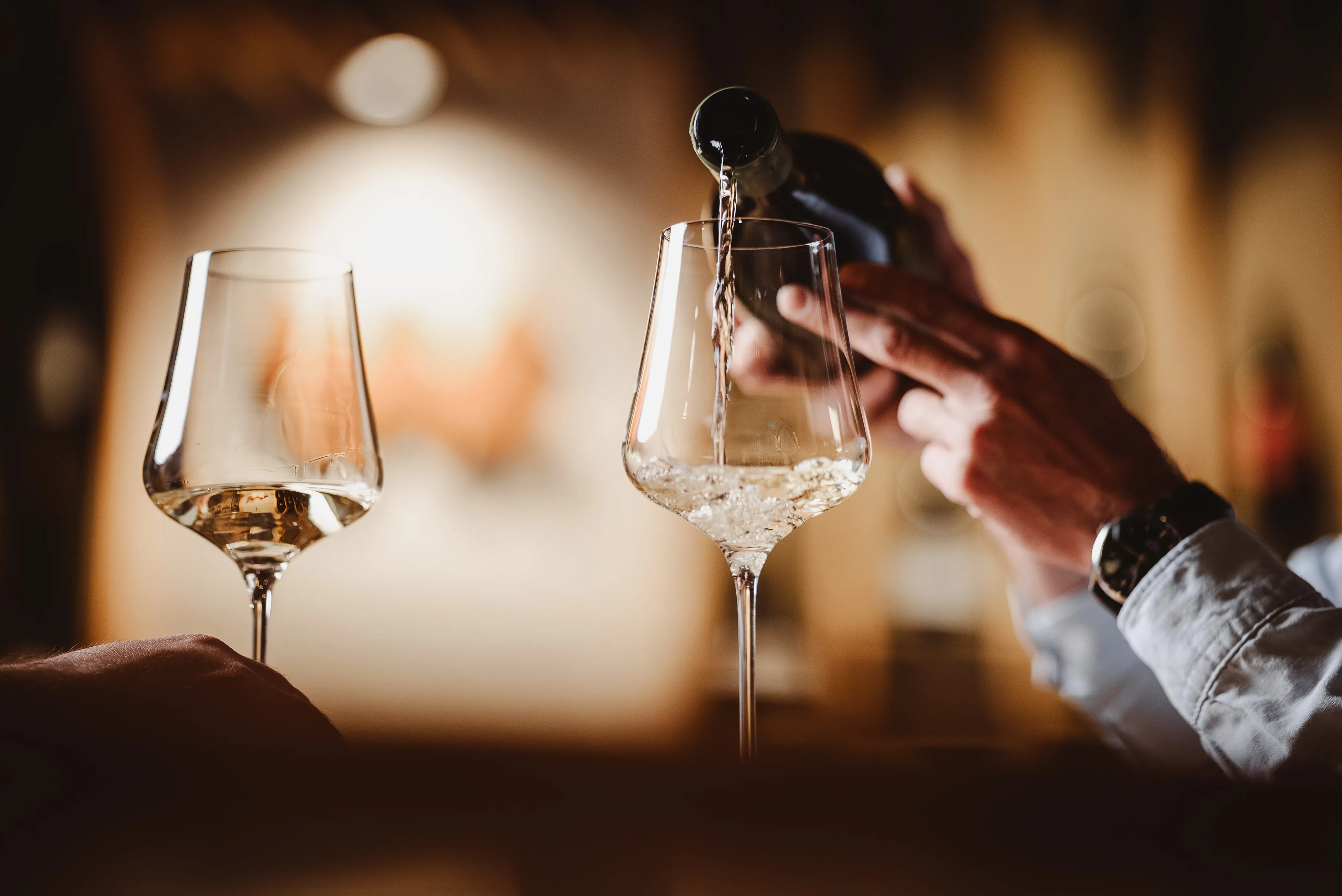 Hand pouring white wine from a bottle into a wine glass next to another partially filled glass.