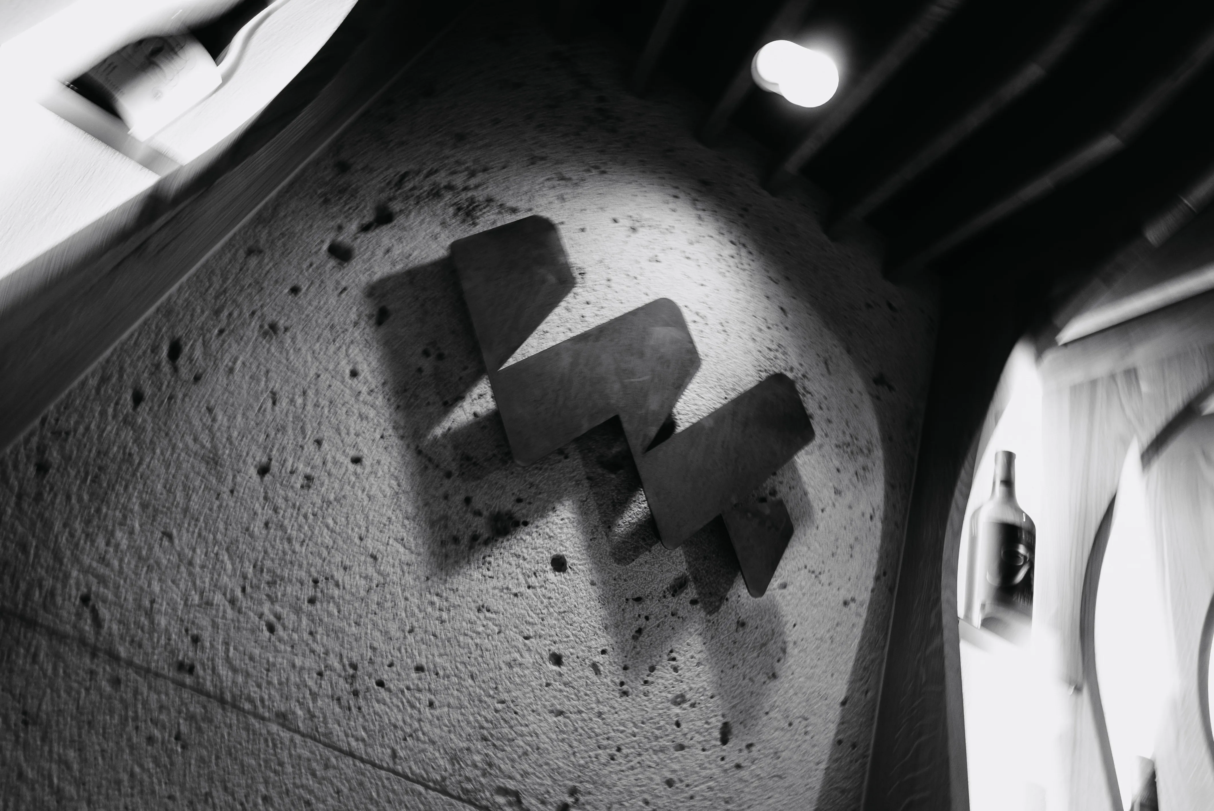 Black metal logo casting shadows on a textured wall under a spotlight in a dimly lit room.