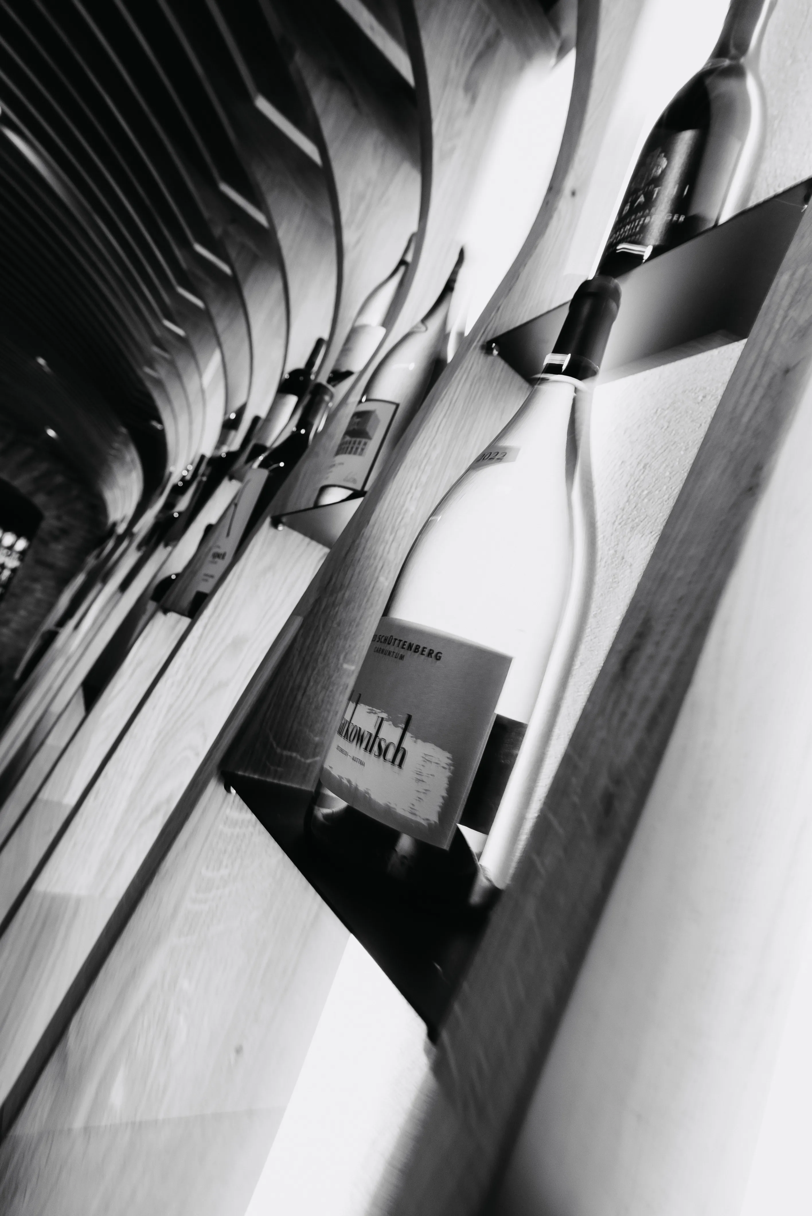 Black and white photo of wine bottles displayed in angled wooden racks along a curved wall.