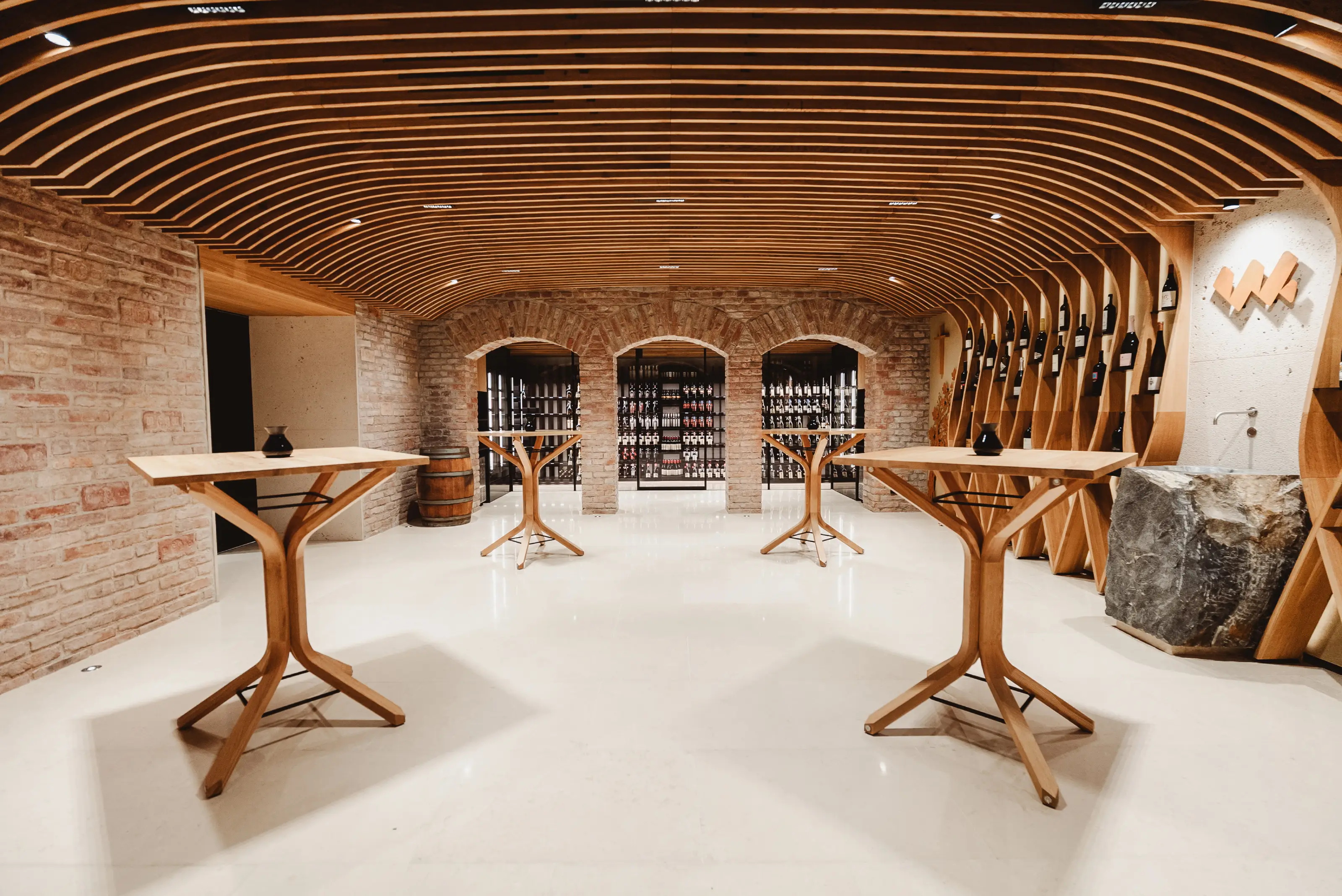 Modern wine cellar with wooden arch ceiling, brick walls, wine bottles on shelves, and four wooden tables.