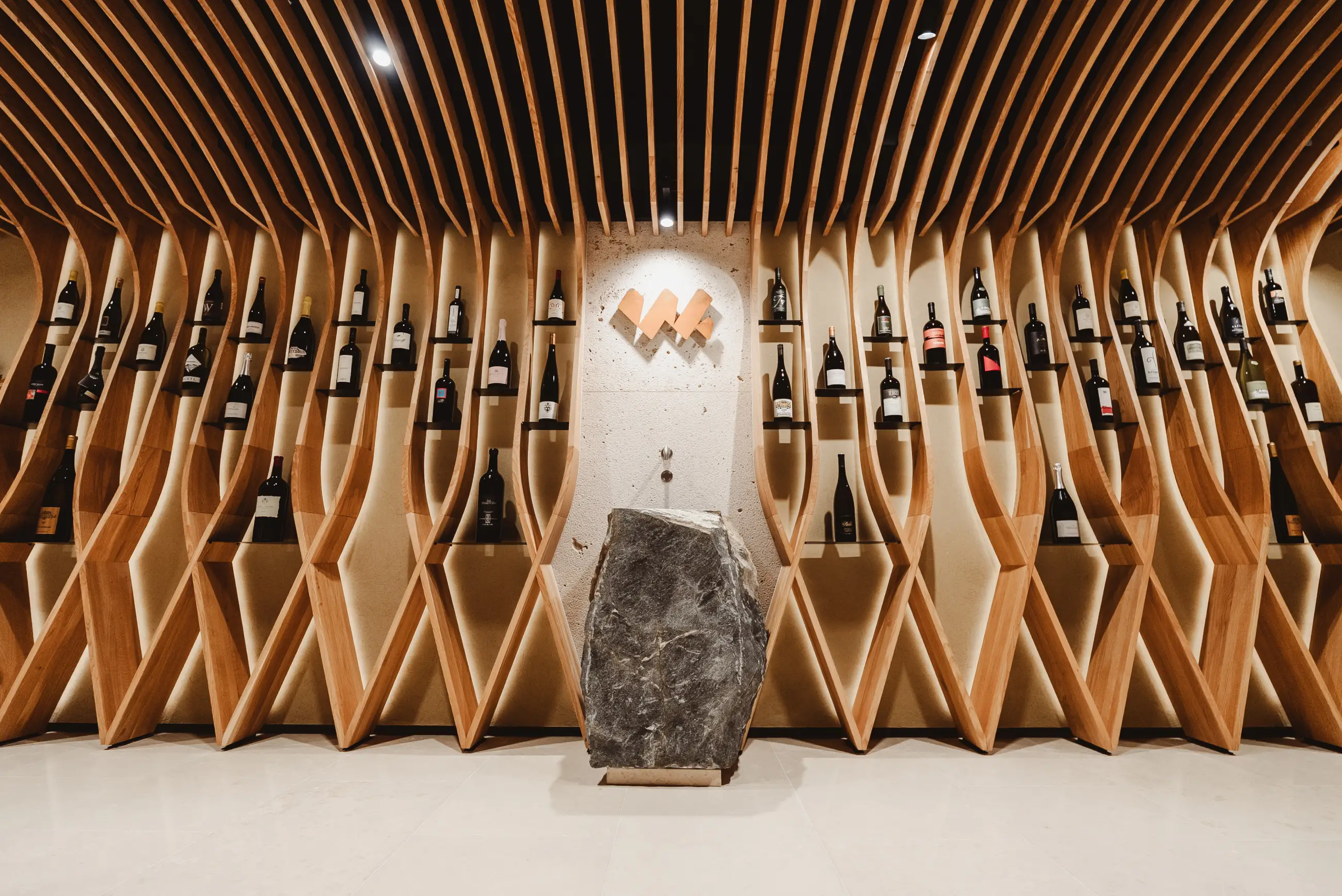 Modern wine cellar with wooden curvy racks holding various wine bottles and a large stone pedestal in the center.