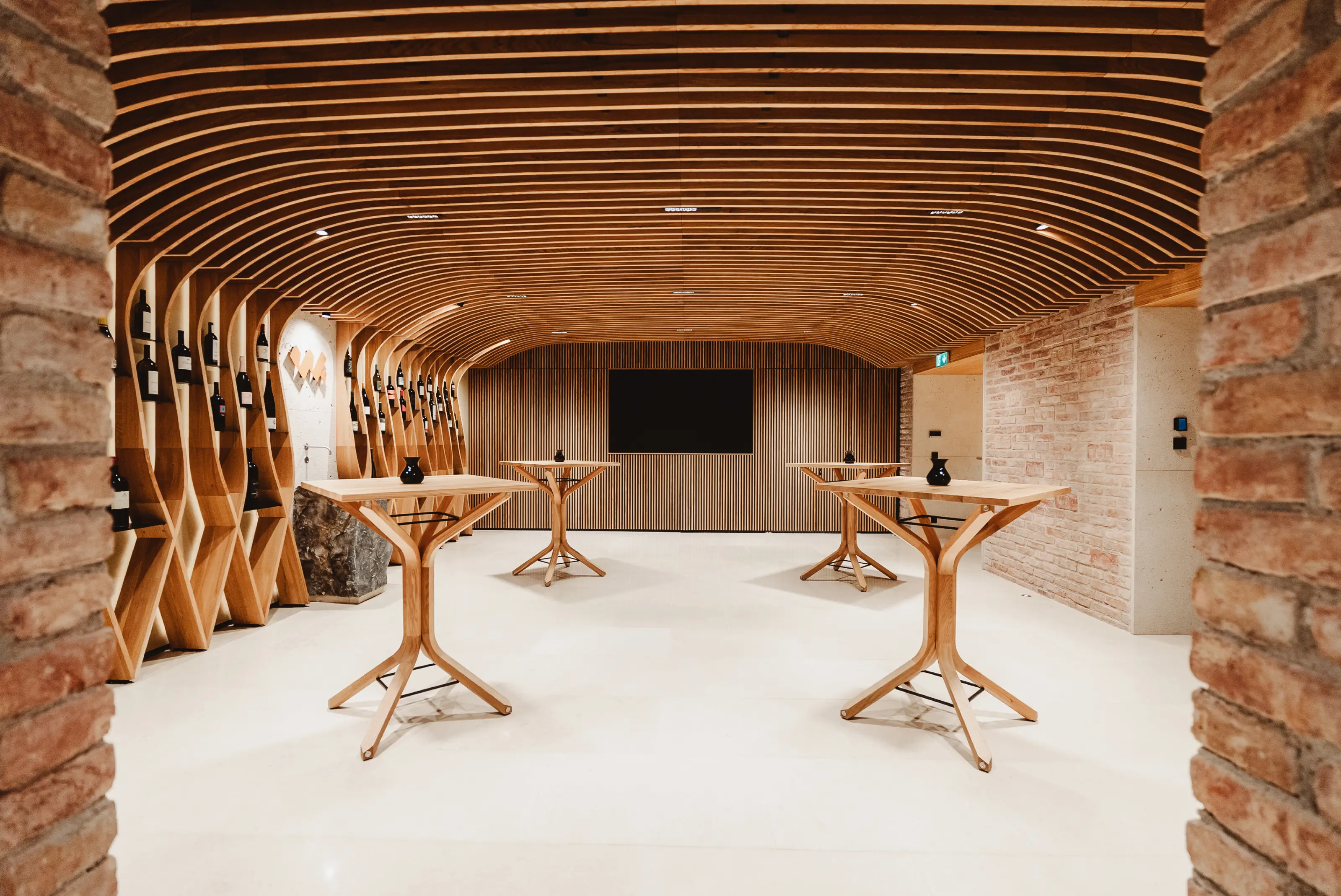 Modern tasting room with curved wooden ceiling, wine bottles on wooden racks, and four high wooden tables.