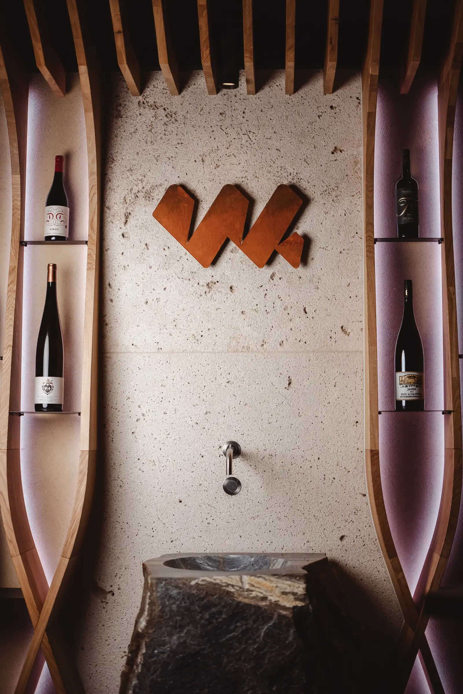 Stone sink with wall-mounted faucet beneath a wooden logo and wine bottles displayed in curved wooden shelves.