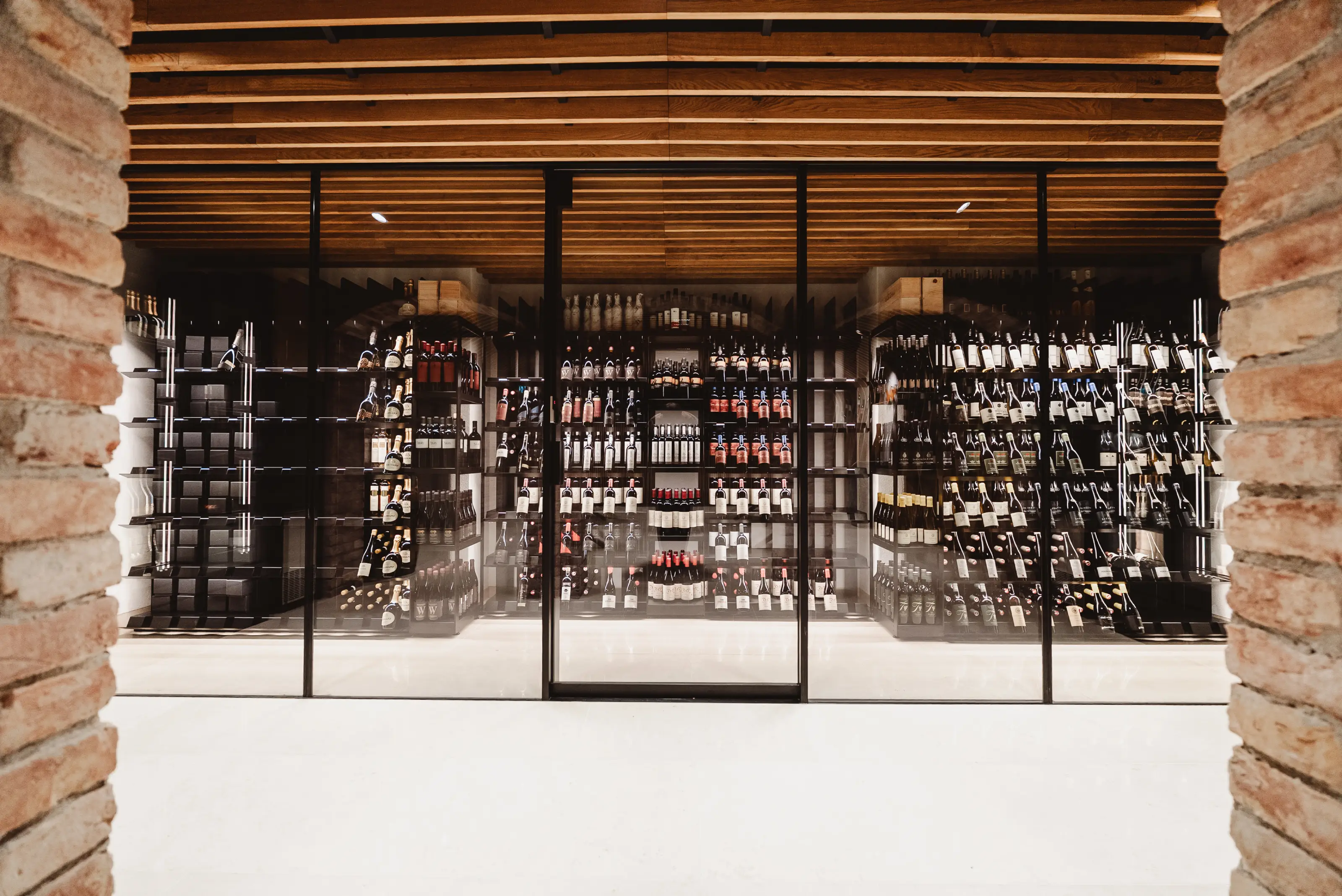 Modern wine cellar with wooden ceiling and glass doors showcasing bottles on black racks.