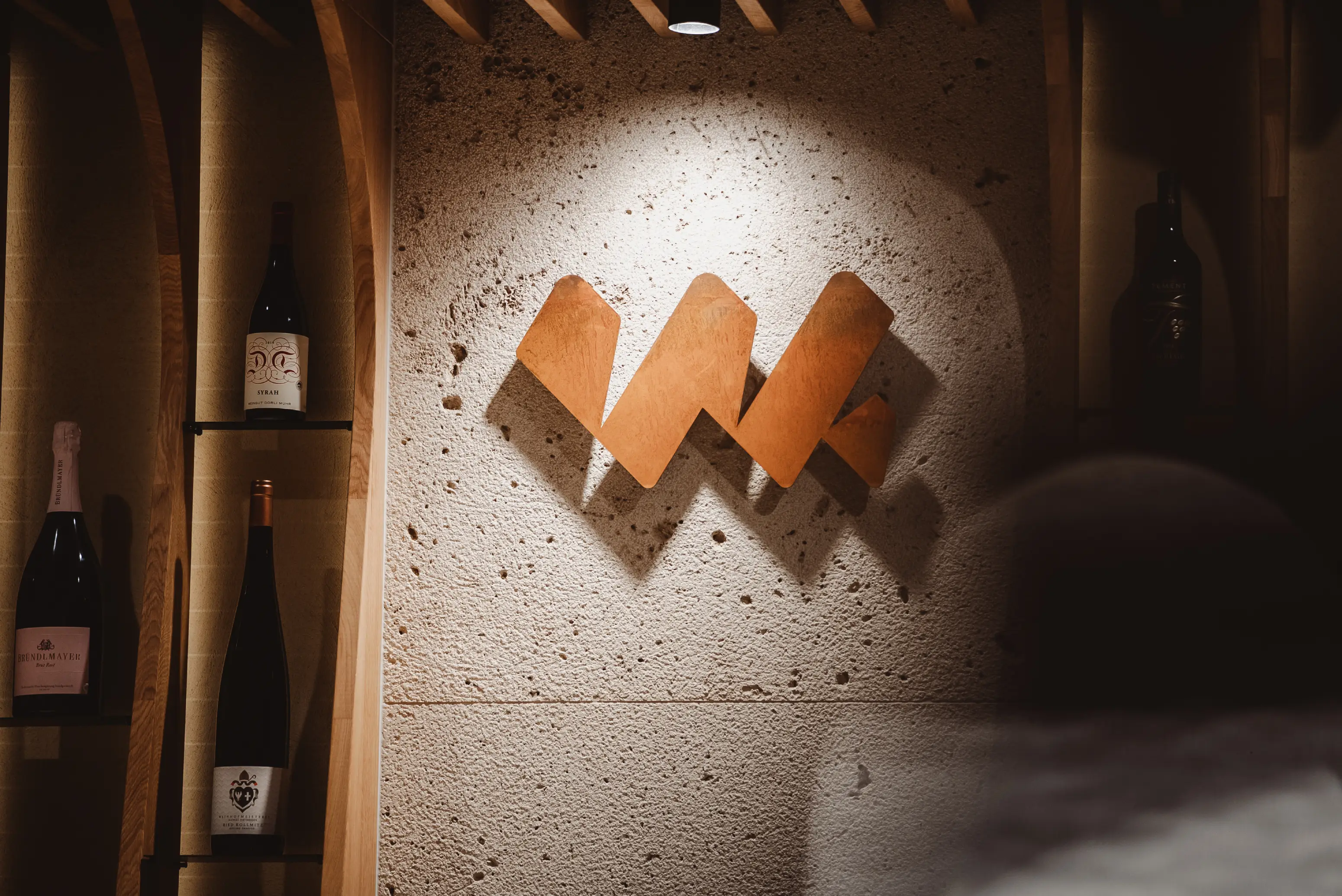 Wine bottles displayed on wooden shelves next to a textured wall with a spotlighted abstract wooden logo.